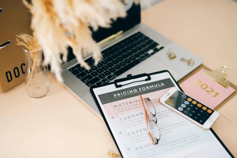 Organized pricing formula document on a desk next to a laptop and smartphone calculator
