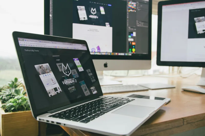 Laptop and monitors on a desk showing software interface used to build and manage digital marketing pages